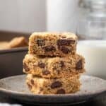 Three chocolate chip cookie bars stacked on a plate with a glass of milk in the background.