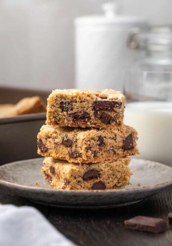 Three chocolate chip cookie bars stacked on a plate with a glass of milk in the background.