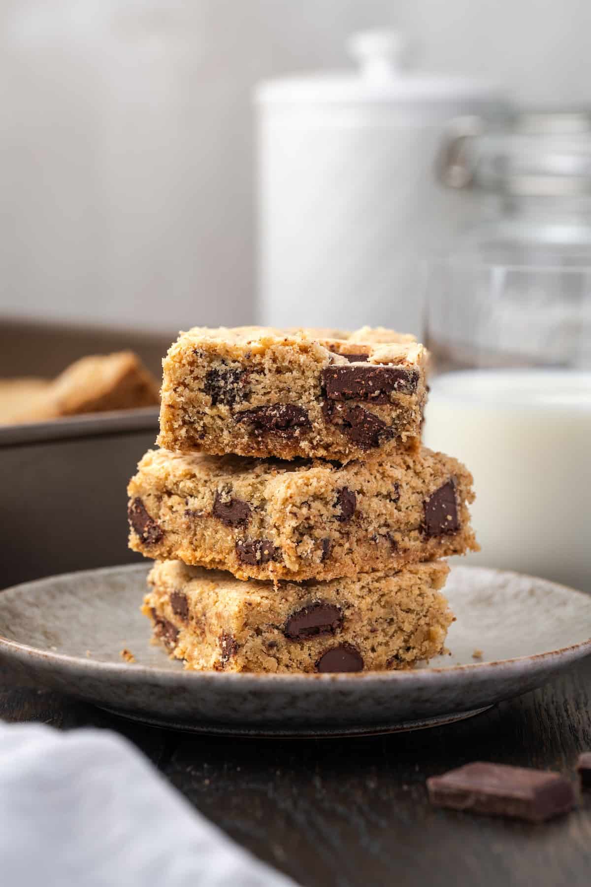 Three chocolate chip cookie bars stacked on a plate with a glass of milk in the background.