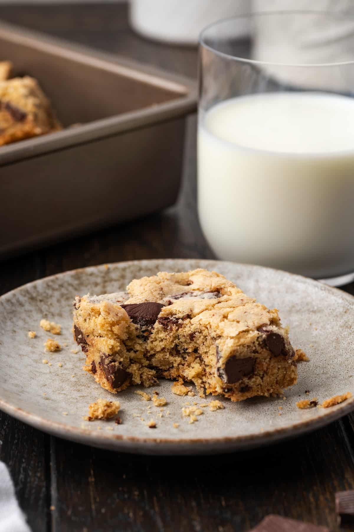 A chocolate chip cookie bar with a bite missing on a plate, with a glass of milk on the background.
