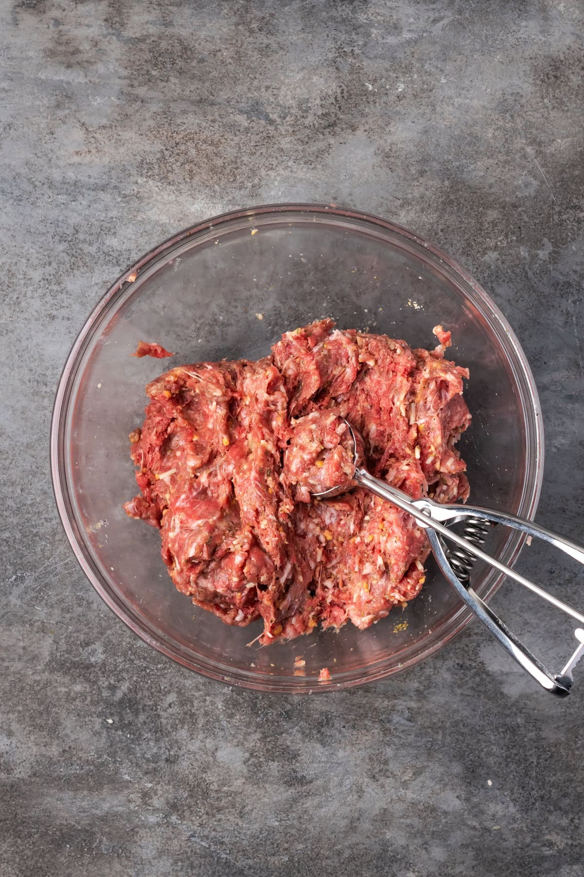 A cookie scoop resting in a bowl of meatball mixture.