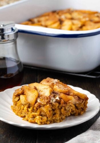A slice of baked pumpkin oatmeal on a plate with the rest of the baked oatmeal in a baking dish in the background.