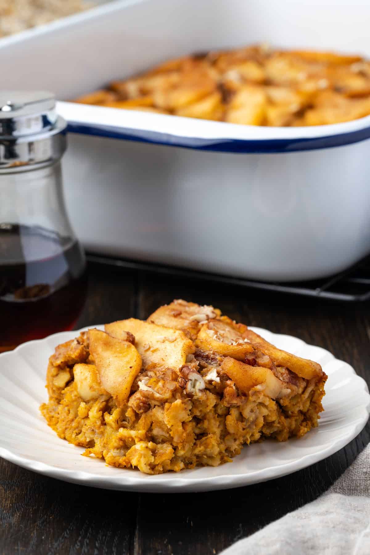 A slice of baked pumpkin oatmeal on a plate with the rest of the baked oatmeal in a baking dish in the background.