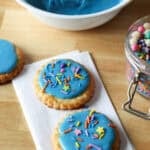Two cookies decorated with blue sugar cookie icing and sprinkles on a countertop, with a bowl of blue icing in the background.