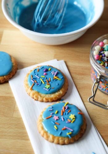 Two cookies decorated with blue sugar cookie icing and sprinkles on a countertop, with a bowl of blue icing in the background.
