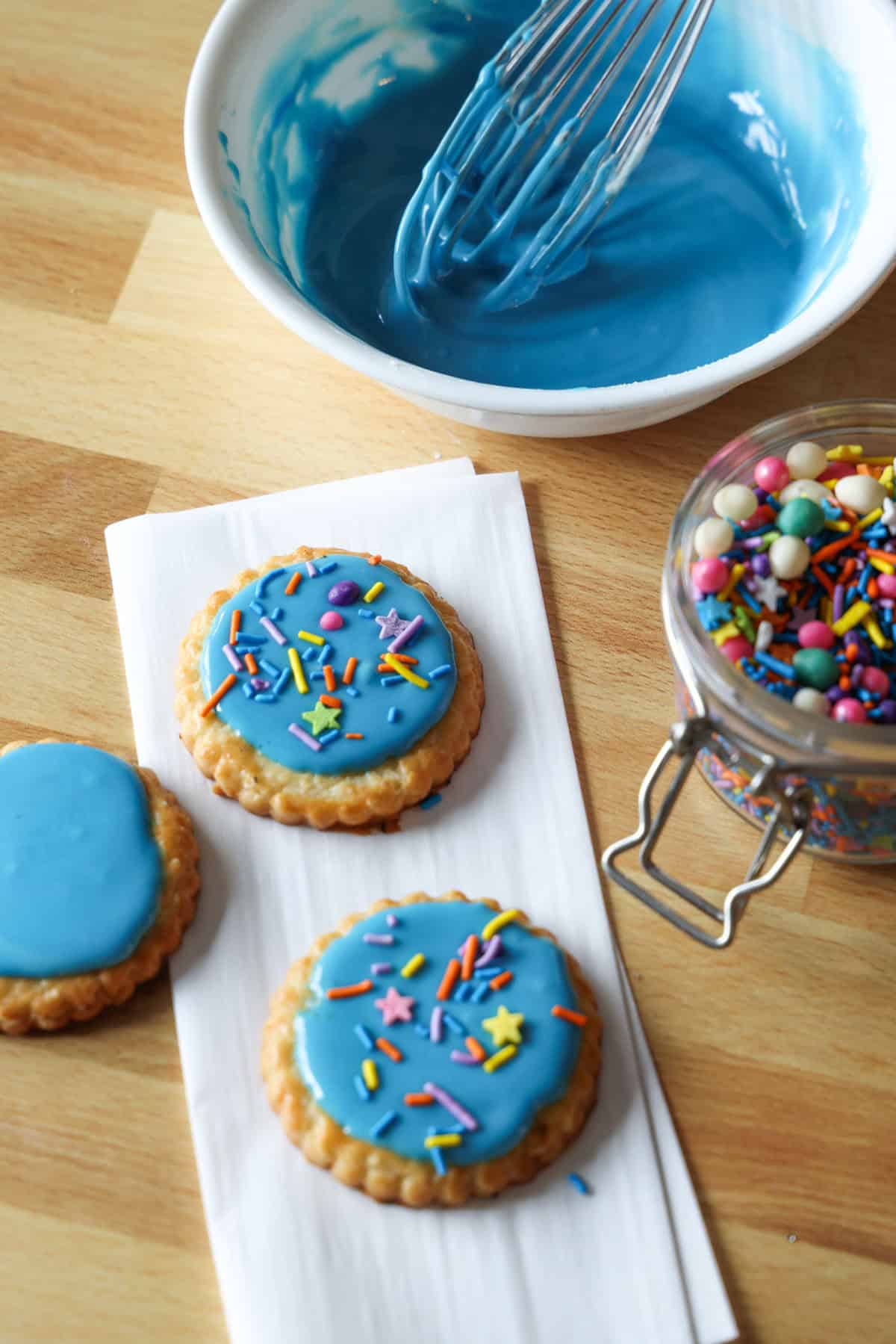 Overhead view of sugar cookies decorated with blue cookie icing and sprinkles on a countertop, next to a bowl of blue icing and a jar of sprinkles.