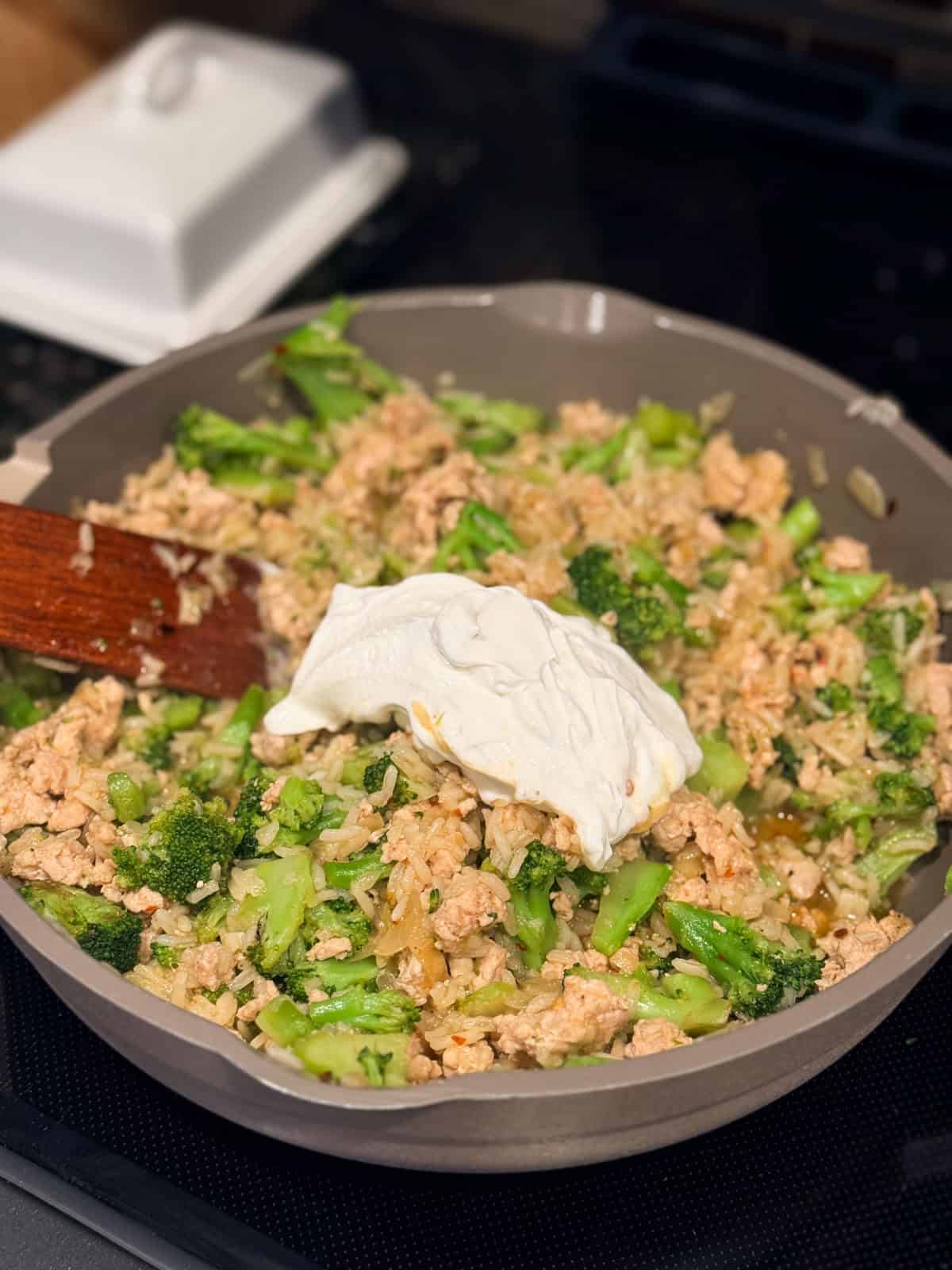 A dollop of sour cream added to a skillet with chicken and broccoli.