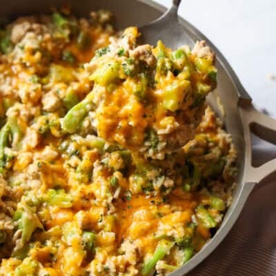 A spoon scooping cheesy chicken and broccoli with rice from a skillet.