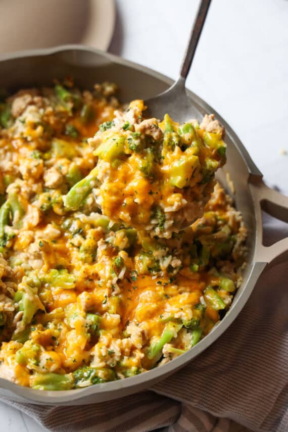 A spoon scooping cheesy chicken and broccoli with rice from a skillet.