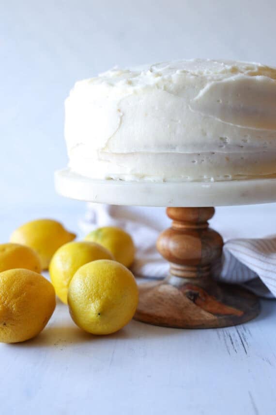 Once the cakes have baked, cool them and then level them off if domes have formed (if necessary). Use a serrated knife to slice the cakes in half. Layered Lemon Cake on a cake stand surrounded by lemons