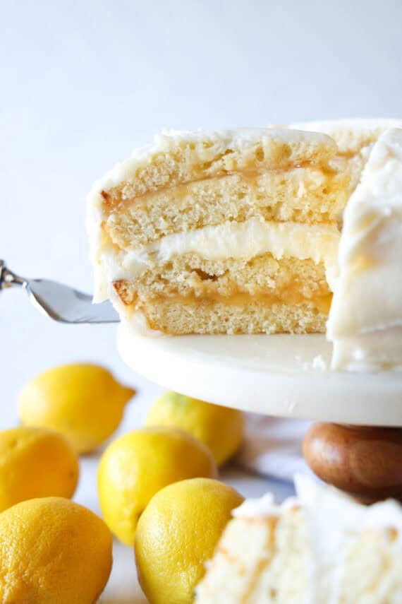 Once the cakes have baked, cool them and then level them off if domes have formed (if necessary). Use a serrated knife to slice the cakes in half. Layered Lemon Cloud Cake with layers of lemon curd and lemon buttercream sliced on a cake stand