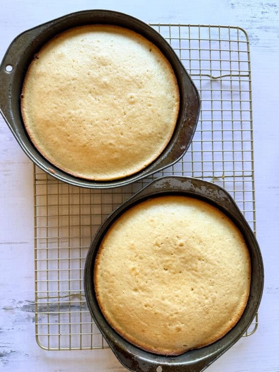 Once the cakes have baked, cool them and then level them off if domes have formed (if necessary). Use a serrated knife to slice the cakes in half. Two baked cakes in pans on a wire cooling rack