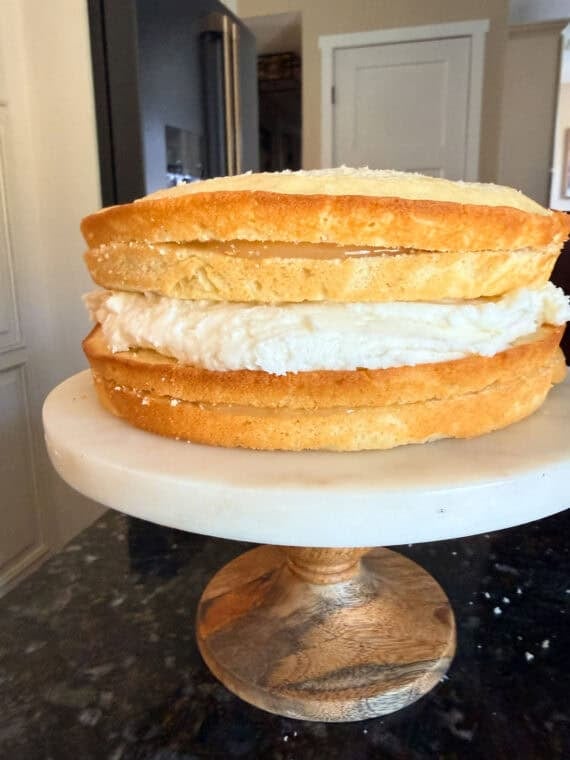 Once the cakes have baked, cool them and then level them off if domes have formed (if necessary). Use a serrated knife to slice the cakes in half. A layered lemon cake on a cake stand showing the filling and layers before frosting the outside.