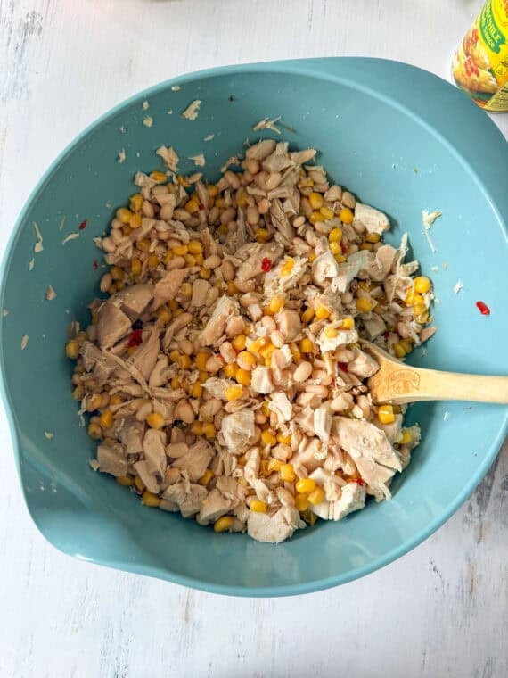 This Chicken Enchilada Casserole is going to earn a regular spot on your menu. It has all the cozy flavors of classic chicken enchiladas layered into an easy casserole Mixing chicken, corn, and beans together in a blue mixing bowl.