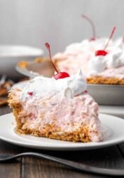 A slice of millionaire pie topped with whipped cream and a cherry on a plate, with the rest of the pie in the background.