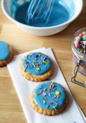 Two cookies decorated with blue sugar cookie icing and sprinkles on a countertop, with a bowl of blue icing in the background.