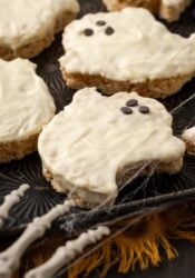 Halloween rice krispie treat ghosts on a baking sheet, decorated with faces made from mini chocolate chips, with a plastic skeleton's fingers in the corner of the frame.