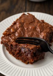 A fork cuts into the corner of a slice of Coca Cola cake on a white plate.