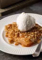 A serving of apple dump cake on a white plate topped with a scoop of ice cream, next to a spoon.